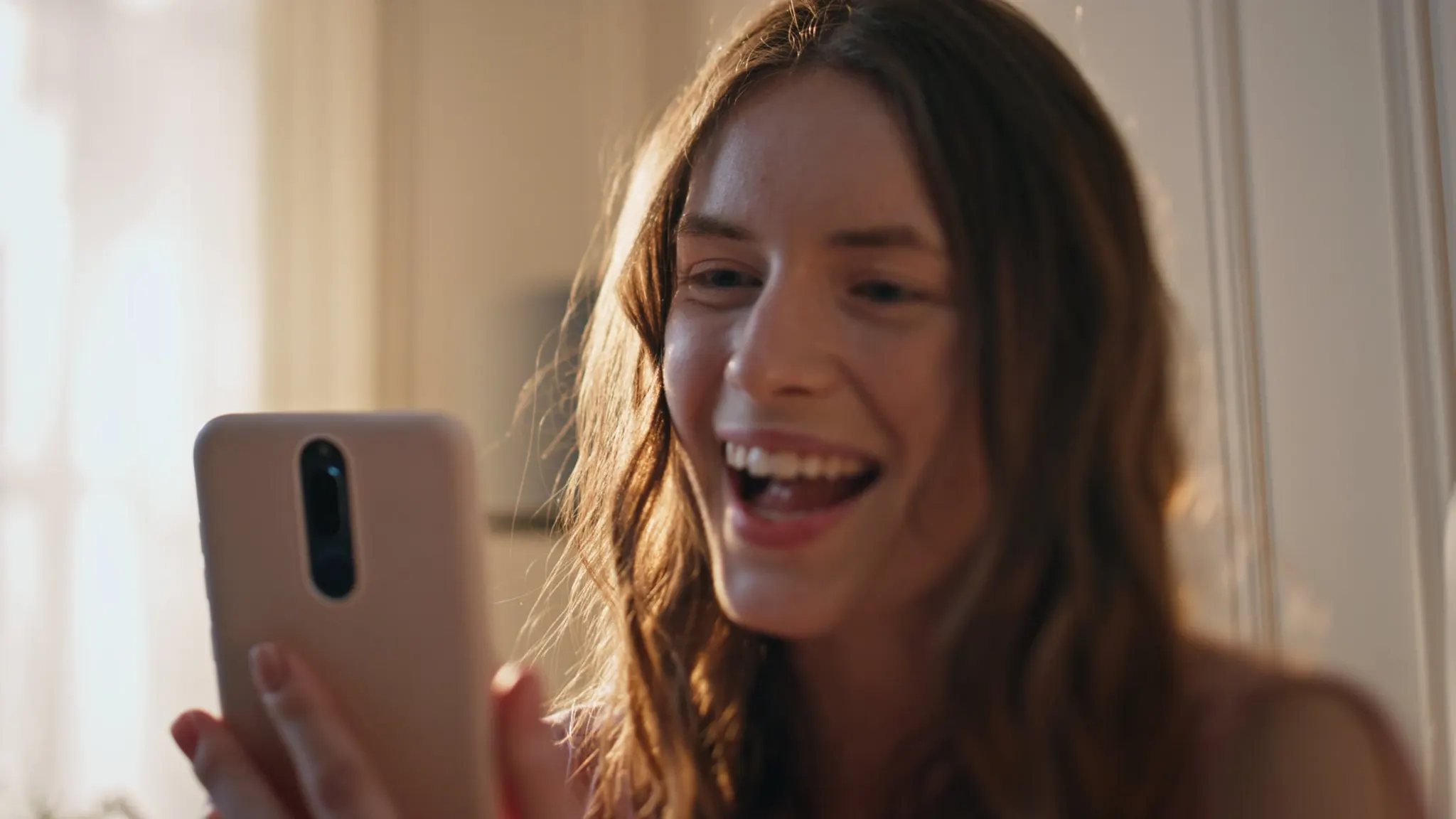 Une femme souriante tient son téléphone dans une chambre baignée de soleil